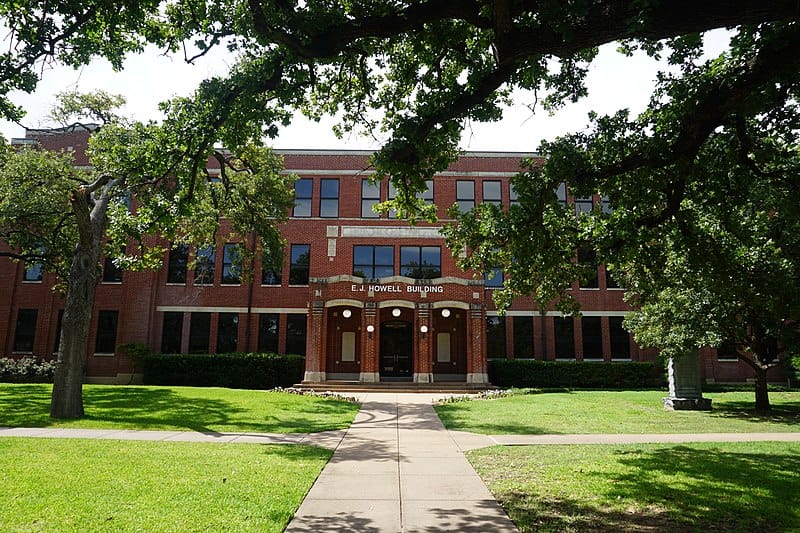 A red brick building labeled 'E.J. Howell Building' is centrally located in the image. It has multiple windows and a prominent entrance with three round lamps. The foreground features a well-maintained lawn with a concrete pathway leading to the entrance. Large trees with green foliage frame the building on both sides, casting shadows on the grass.
