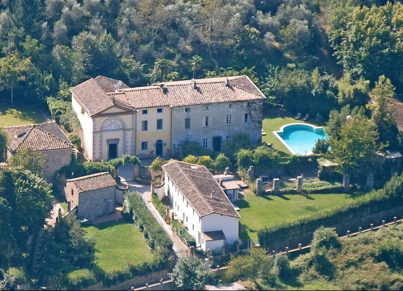 An aerial photograph of a large Italian villa surrounded by lush greenery and trees. The villa features traditional architecture with a tiled roof and several buildings. There is a well-maintained garden and a rectangular swimming pool adjacent to the main building. The property is enclosed by hedges and is set against a backdrop of dense forest.