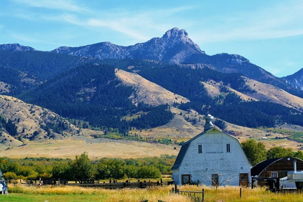 A picturesque rural scene featuring a white barn with a gray roof situated in a grassy field. In the background, there are majestic mountains with a mix of rocky and forested areas under a clear blue sky. The foreground includes fenced pastures and scattered trees, adding to the idyllic countryside setting.