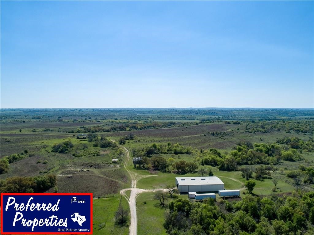 This image captures an expansive rural landscape under a clear blue sky. In the foreground, there is a large, rectangular white building with a few smaller structures nearby, set amidst green fields and scattered trees. A dirt road leads to the buildings, winding through the verdant terrain. The background showcases a vast expanse of rolling hills and open fields extending towards the horizon.