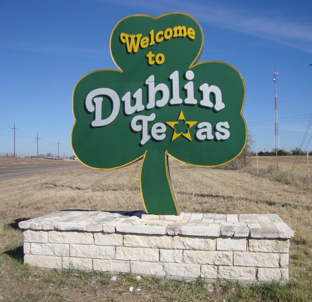 A large green shamrock-shaped sign with yellow and white lettering welcomes visitors to Dublin, Texas. The sign is mounted on a stone base and is situated in a rural area with open fields and a clear blue sky. Telephone poles and a communication tower are visible in the background.