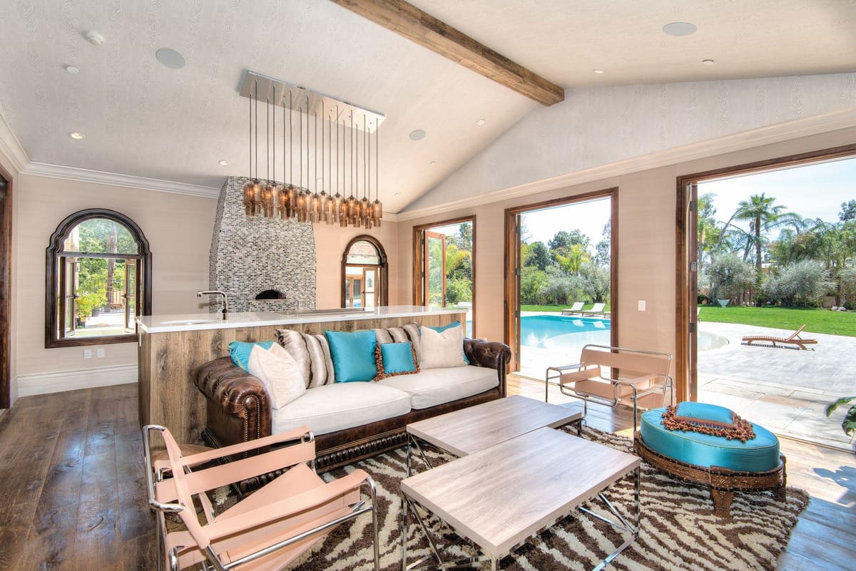 A spacious and elegant living room featuring a large leather and fabric sofa adorned with turquoise and beige pillows. The room has a high ceiling with exposed wooden beams and a modern chandelier with hanging lights. A tiled fireplace with a mosaic pattern is a focal point. Large glass doors open to a view of a swimming pool and lush greenery outside. The room includes contemporary chairs and a zebra-patterned rug, creating a stylish and comfortable atmosphere.