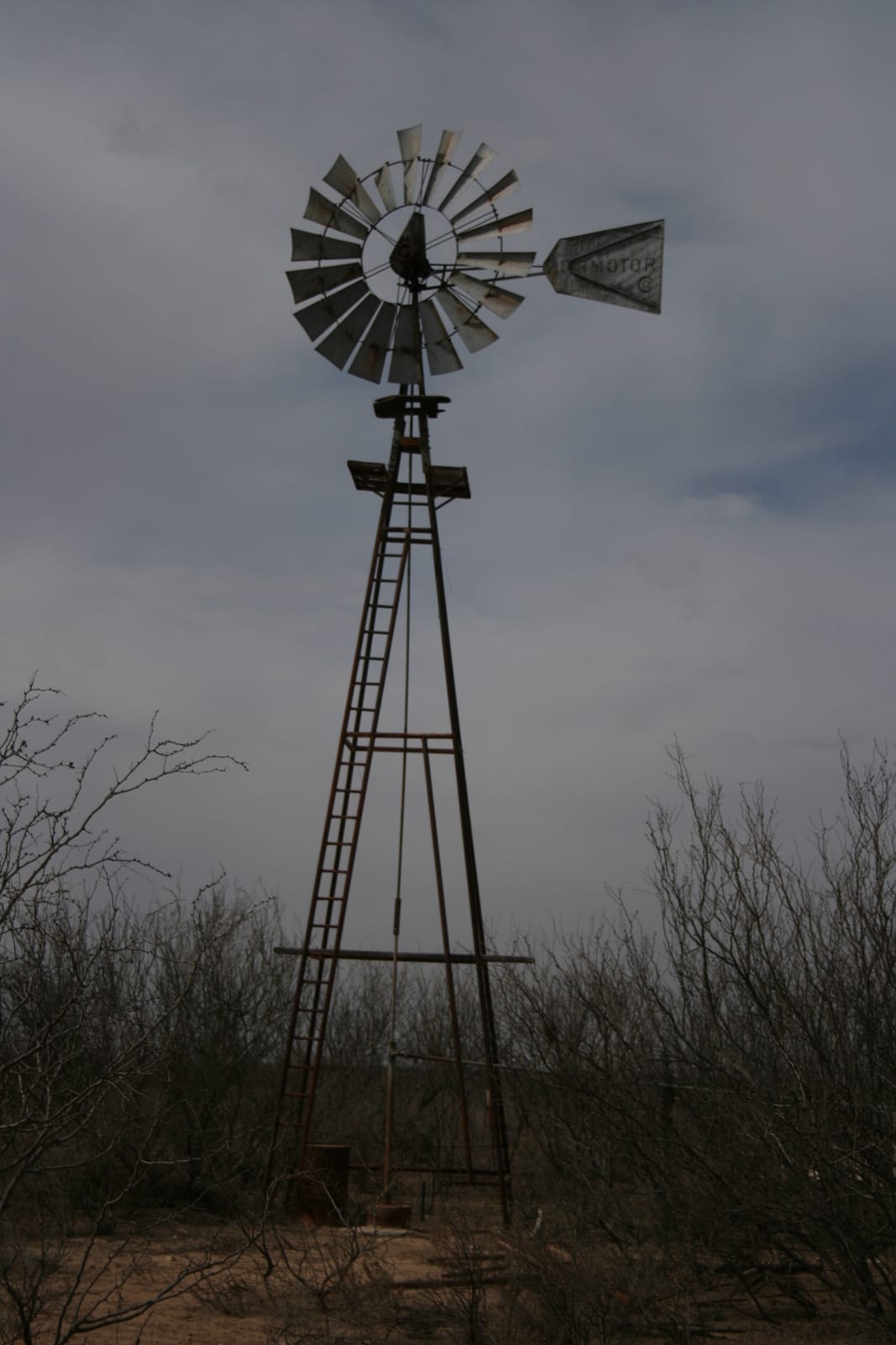A tall, metallic windmill stands against a cloudy sky. The windmill has a classic design with a circular rotor and tail vane. The structure is surrounded by dry, leafless shrubs, suggesting an arid or semi-arid environment. The ground appears barren, with patches of dry soil visible. The overall atmosphere is calm and slightly overcast.