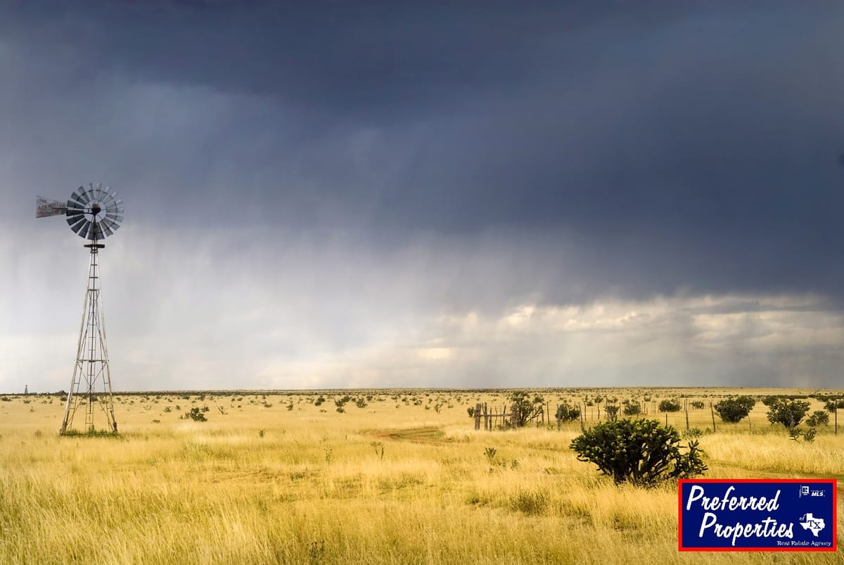 A solitary windmill stands in a vast, open grassland under a dramatic, cloudy sky. The landscape is flat with sparse vegetation, and the grass is golden brown, suggesting a dry environment. A storm appears to be approaching, with dark, heavy clouds dominating the upper part of the image. On the right side, there is a logo for 'Preferred Properties' with additional text indicating a real estate agency.
