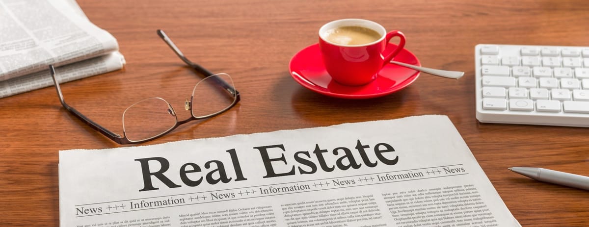 A wooden desk features a newspaper with the headline 'Real Estate' prominently displayed. Nearby, a pair of reading glasses rests on the desk. A red coffee cup with a saucer containing a cappuccino is positioned next to a white wireless keyboard. A pen is also visible on the desk, suggesting a workspace or reading environment.
