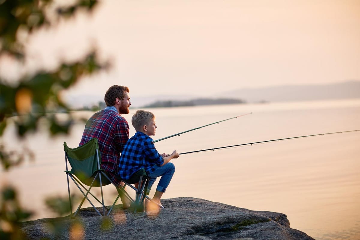 A man and a boy are sitting on foldable chairs on a rocky shore, fishing in a calm lake during sunset. They are both holding fishing rods and wearing plaid shirts, with the man in a red plaid shirt and the boy in a blue one. The background features a serene lake with gentle ripples and distant hills under a pinkish sky. The scene is peaceful, with some blurred foliage in the foreground adding a natural frame.