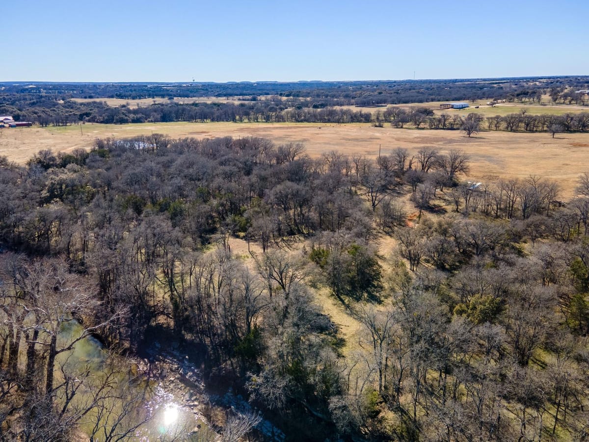 An aerial photograph showcasing a large expanse of land featuring a dense forest area in the foreground, transitioning into open fields in the background. The forest is composed of leafless trees, indicating a winter or early spring setting. A small body of water is visible at the bottom left, reflecting sunlight. The horizon is visible, with distant hills and a clear blue sky. Sparse buildings are scattered across the landscape.