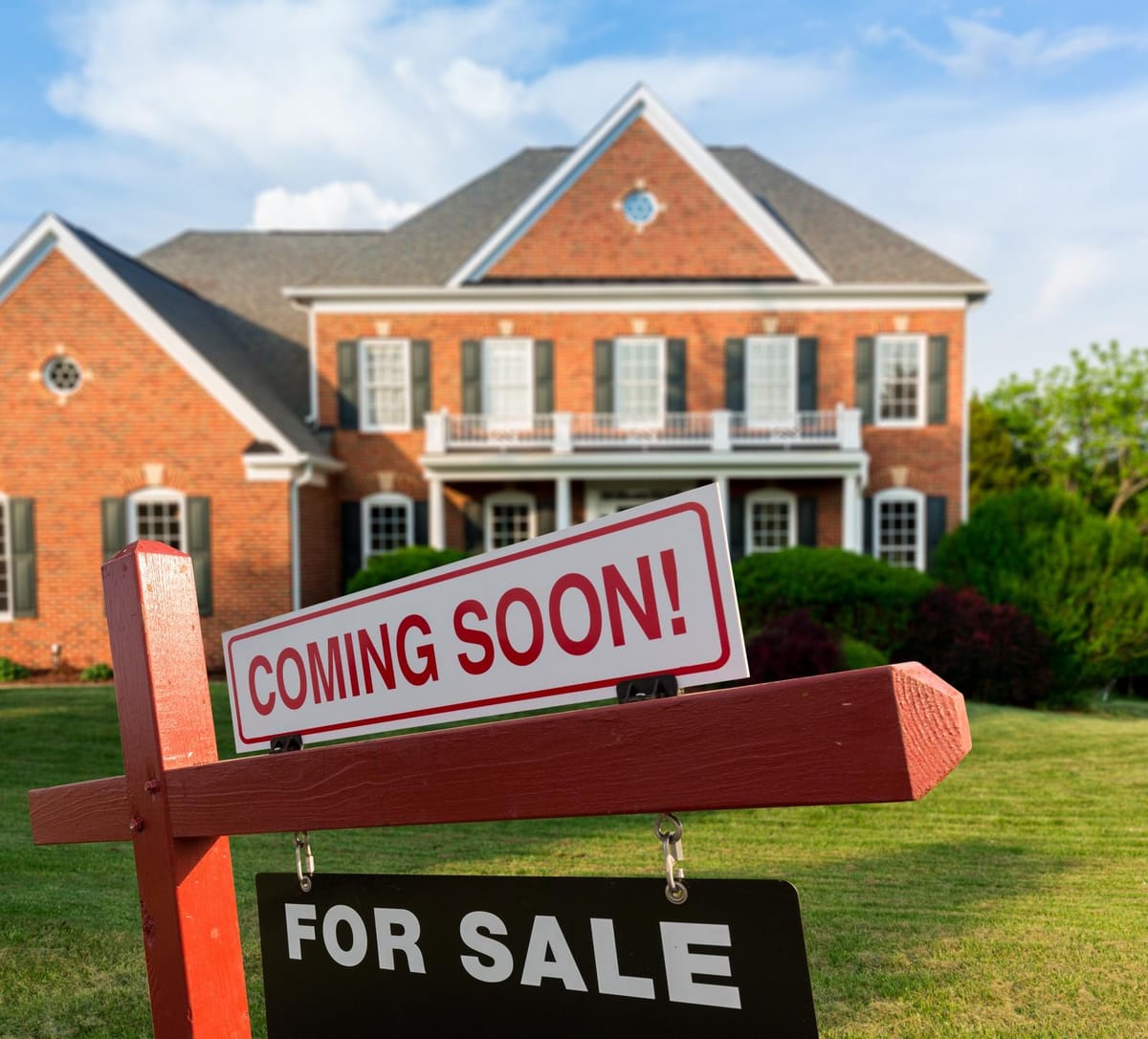 A large brick house with a symmetrical facade, featuring multiple windows with shutters and a small balcony. The house is set in a well-maintained yard with green grass and bushes. In the foreground, there is a wooden post with two signs attached: a red and white 'COMING SOON!' sign above a black 'FOR SALE' sign. The sky is clear with a few clouds, suggesting a sunny day.
