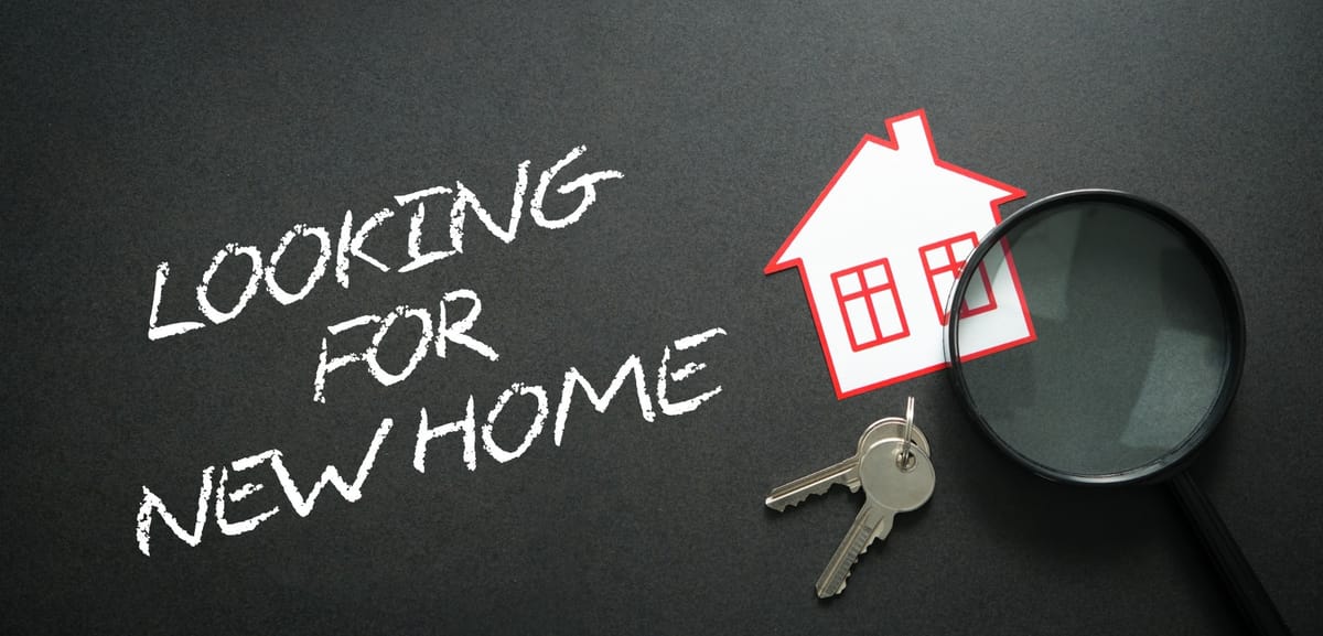 The image features a dark background with the phrase 'Looking for New Home' written in white chalk-like font. To the right, there is a paper cutout of a house with red outlines, a pair of silver keys, and a magnifying glass. The composition suggests themes of real estate, home searching, and property investment.