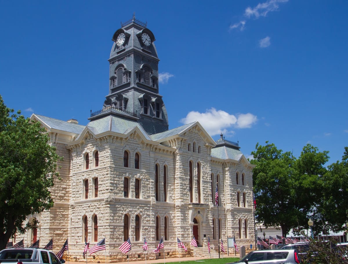 A historic courthouse building with a prominent clock tower under a clear blue sky. The structure is made of light-colored stone with tall arched windows. The clock tower features four clock faces and intricate architectural details. American flags line the perimeter of the building, suggesting a patriotic theme. Trees and parked cars are visible in the foreground, adding to the setting of a civic area.