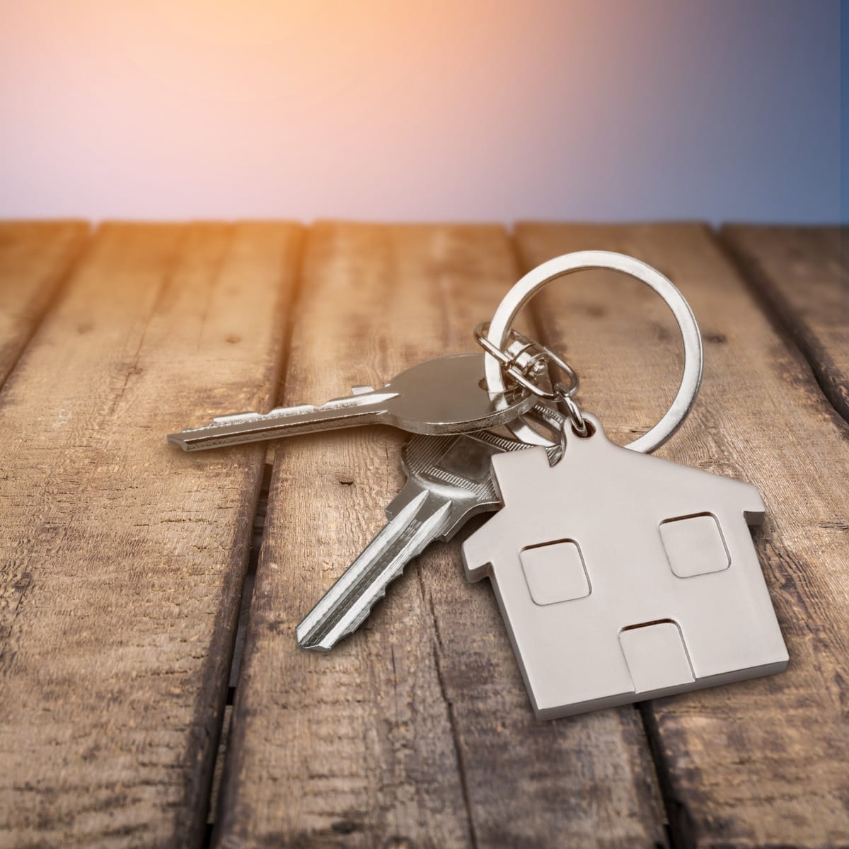 A set of metallic keys attached to a house-shaped keychain lies on a wooden surface. The keychain is silver in color and features simple cutouts representing windows and a door. The wooden surface consists of planks with visible grain and texture. The background has a gradient from warm orange to soft blue, suggesting a sunrise or sunset ambiance.