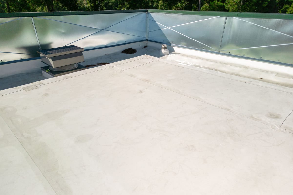The image shows a flat rooftop with a light-colored surface. There is a ventilation unit positioned near the corner of the roof. The perimeter of the roof is enclosed with a metallic railing featuring diagonal support bars. The background shows a view of trees, indicating an outdoor setting. The rooftop appears clean and well-maintained.