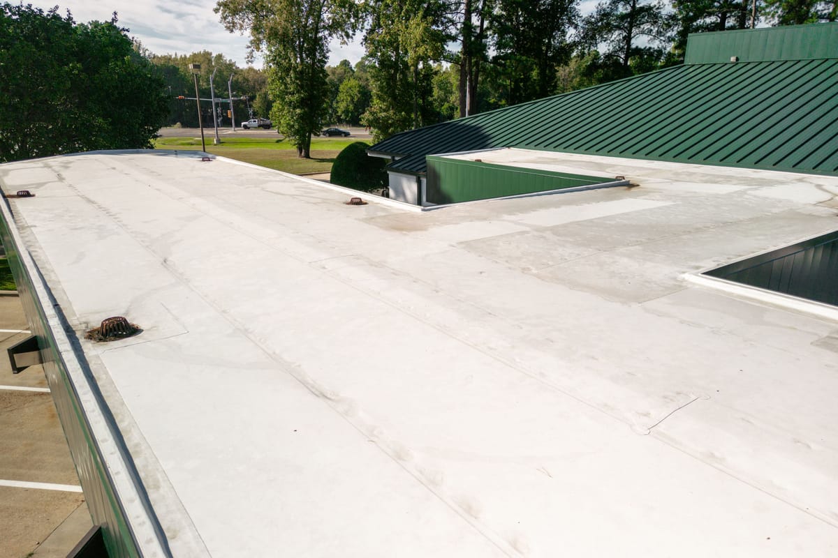 The image shows a flat commercial roof with a white surface and visible drainage systems. The roof is part of a building with a green metal siding and roof section. Trees and a road with cars are visible in the background, indicating an urban or suburban setting. The roof appears to be well-maintained with minimal debris.