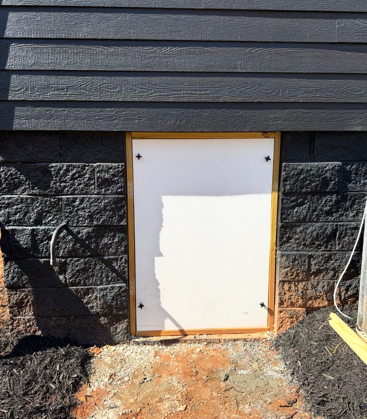 Small Rectangular White Access Door Set into Dark Textured Wall with Mulched Ground and Exposed Soil