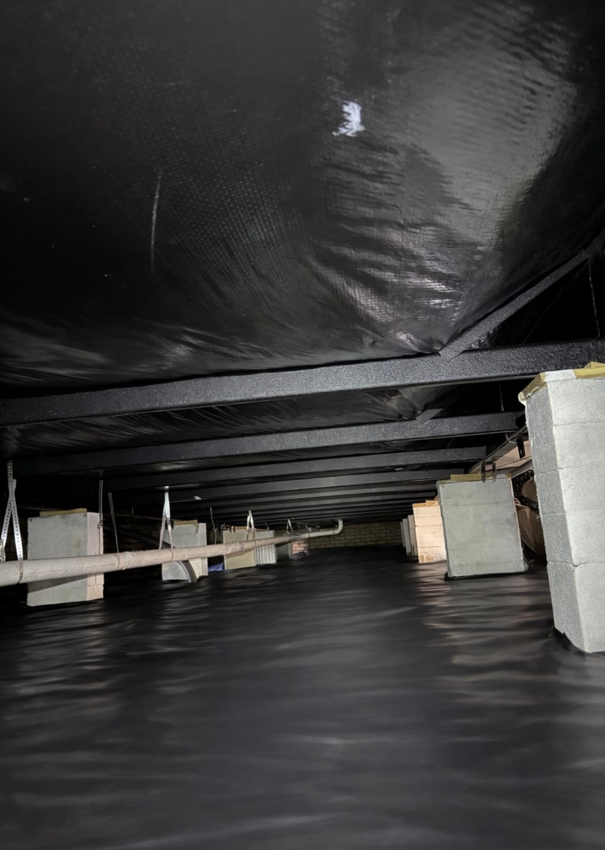 Modern Crawl Space with Black Vapor Barrier and Structural Supports Featuring Exposed Pipes and Concrete Block Piers