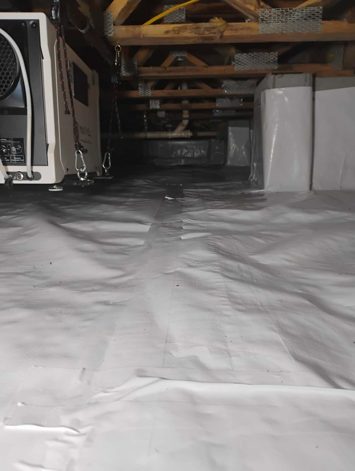 The image depicts a crawl space with a white vapor barrier covering the floor and extending partially up the walls. Wooden beams are visible on the ceiling, supporting the structure. On the left side, a dehumidifier is mounted, suspended by hooks and ropes. The space is clean and organized, suggesting it is well-maintained for moisture control.