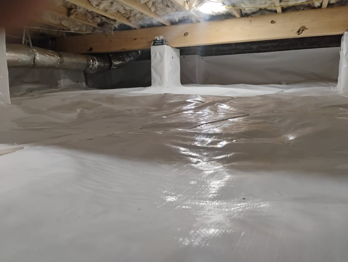 The image shows a crawl space with a newly installed white vapor barrier covering the ground and walls. The ceiling is insulated with exposed wooden beams and insulation material. A metallic duct runs horizontally along the ceiling. The space is well-sealed, with the vapor barrier providing a clean and bright appearance, reflecting light from an unseen source.