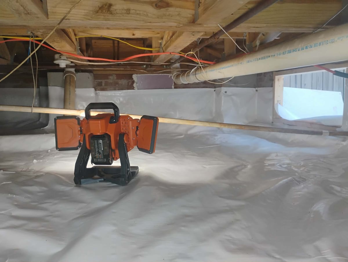 The image shows a crawl space with exposed wooden beams and pipes running along the ceiling. A bright orange portable work light is set up on the floor, illuminating the area. The floor is covered with a white plastic sheeting, likely for moisture protection. Electrical wires are visible running along the beams, and a small window or opening is seen in the background, providing natural light.