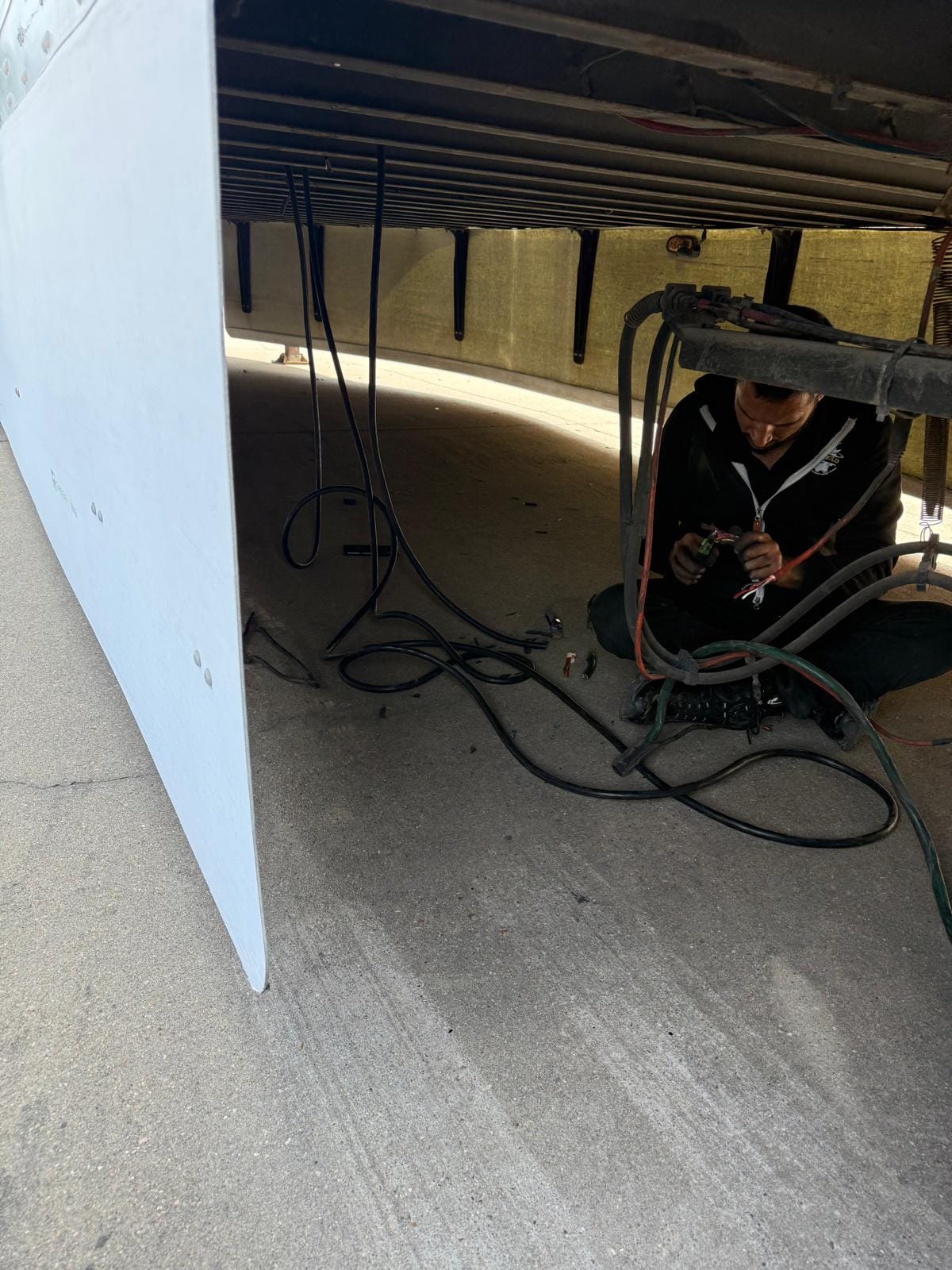 Technician Performing Maintenance Underneath a Large Vehicle with Wiring and Tools on Concrete Surface