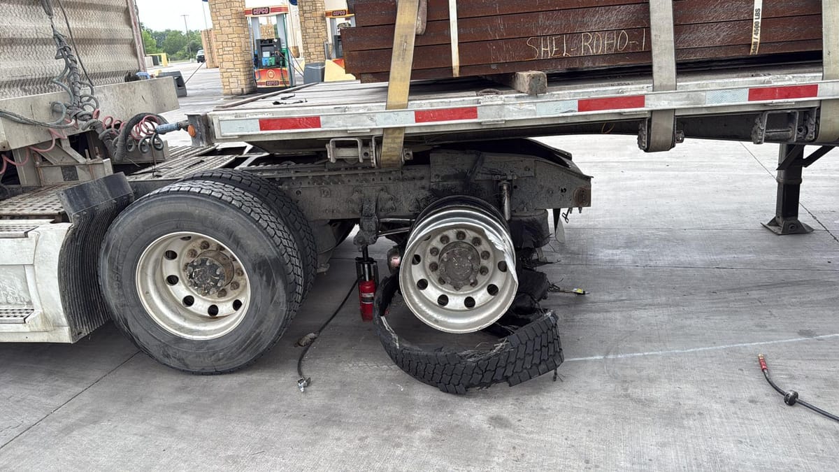 Damaged Rear Wheel of a Semi-Truck at Fuel Station with Exposed Axle and Strapped Metal Cargo