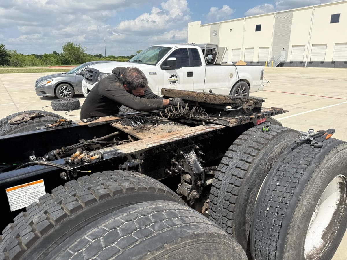 Mechanic Repairing Large Truck Chassis in Open Parking Lot with Multiple Vehicles and Warehouse in Background Under Clear Blue Sky