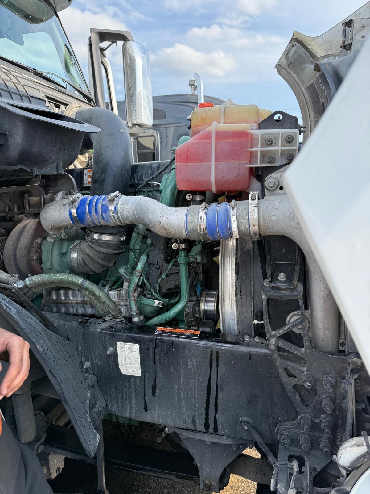 Open Hood of a Heavy-Duty Truck Revealing Engine Components and Coolant Reservoir with Blue and Silver Piping Under a Cloudy Sky
