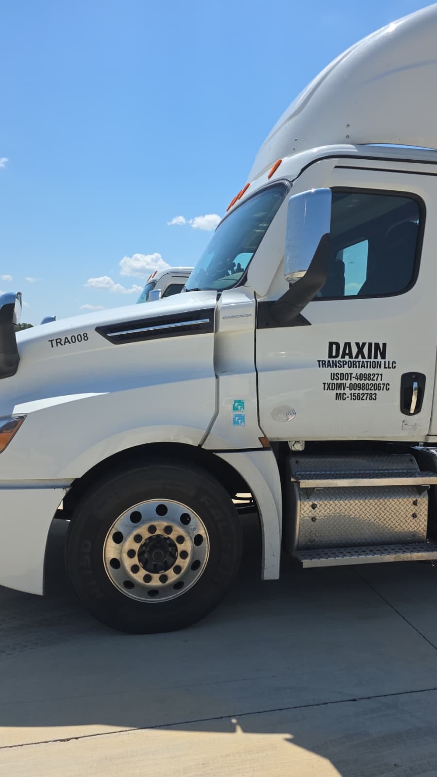 The image shows a white semi-truck parked on a concrete surface against a clear blue sky. The truck has visible branding for 'DAXIN TRANSPORTATION LLC' on the door, along with various identification numbers. The front wheel and part of the cab are visible, featuring chrome elements and a side mirror. The truck's model appears to be a Cascadia, as indicated by the badge near the door.