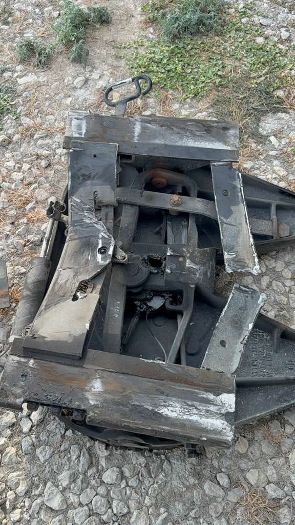 A heavily damaged industrial metal component lies on a gravel surface. The object appears to be part of a larger machinery, showing signs of wear and tear with rust and scratches visible. The surrounding area is covered in small rocks and sparse vegetation, indicating an outdoor setting. The metal structure includes various mechanical parts, bolts, and a handle-like piece.