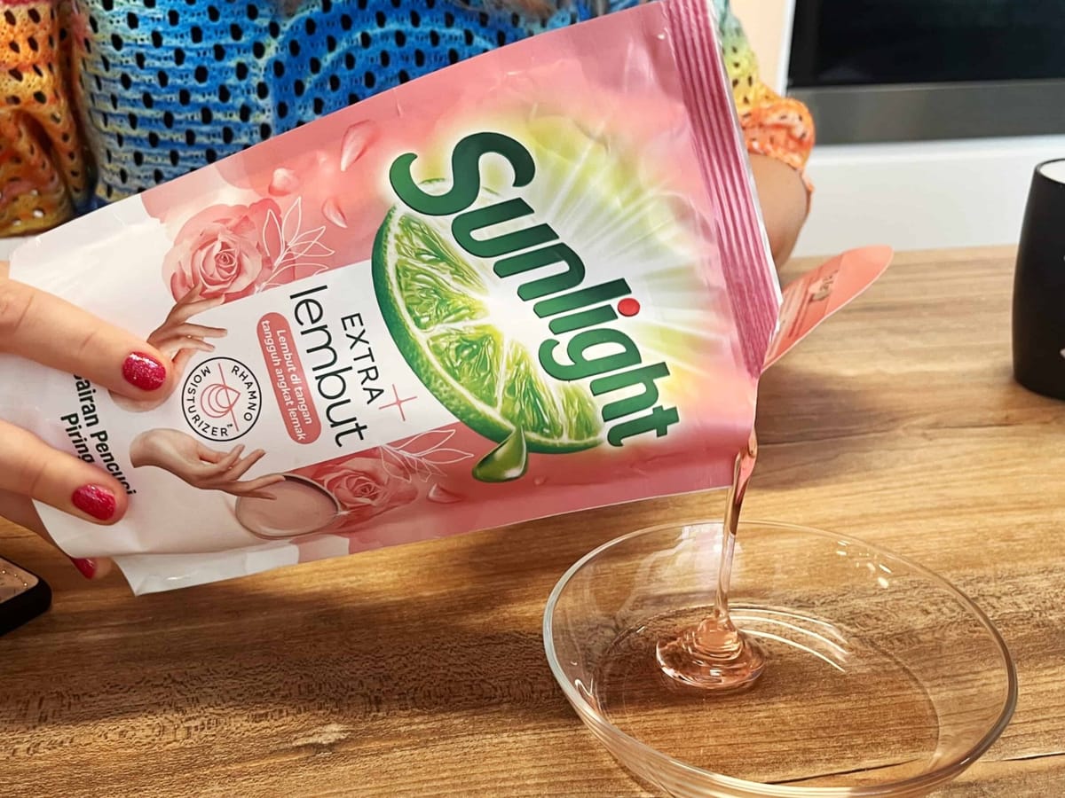 Pouring Pink Liquid Dish Soap from Sunlight Packaging into Clear Glass Bowl on Wooden Table with Colorful Knit Sweater in Background