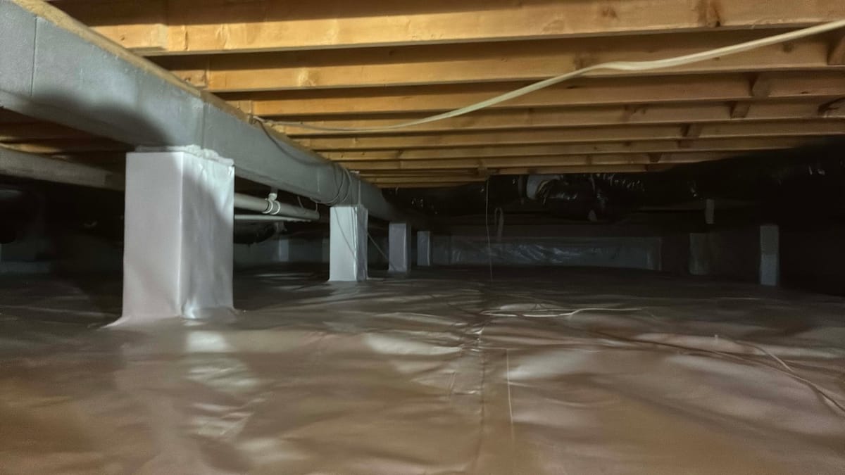 The image depicts a crawl space with a vapor barrier covering the ground. Wooden beams form the ceiling structure, and several support pillars are wrapped in a protective material. Ductwork and piping run along the ceiling, and some wiring is visible. The space is dimly lit, with shadows in the background.