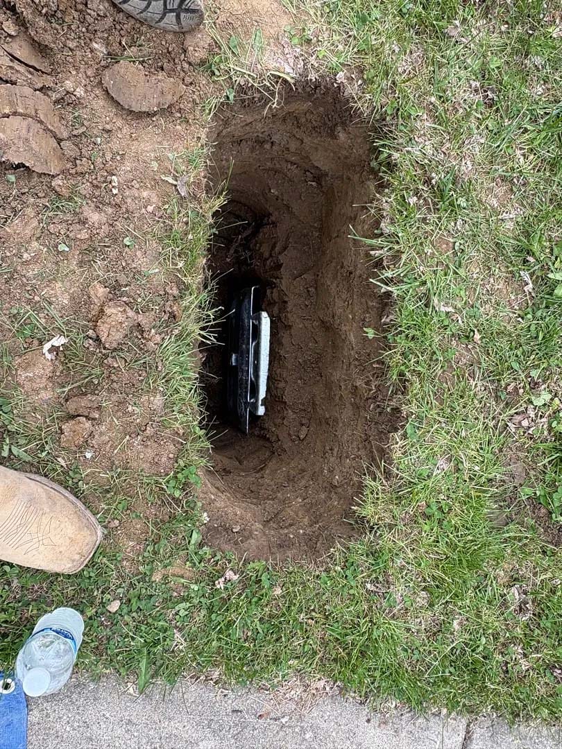 The image shows a shallow hole dug into a grassy area with a partially buried object inside. The object appears to be metallic or plastic. Surrounding the hole, the grass is slightly disturbed, and there is loose soil around the edges. Two shoes are partially visible at the top and bottom left corners, suggesting human presence. A plastic water bottle is placed on the ground near the hole.