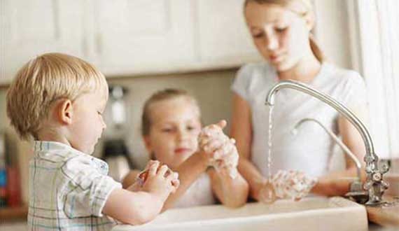 Three Young Children Washing Hands Together at Kitchen Sink with Running Water