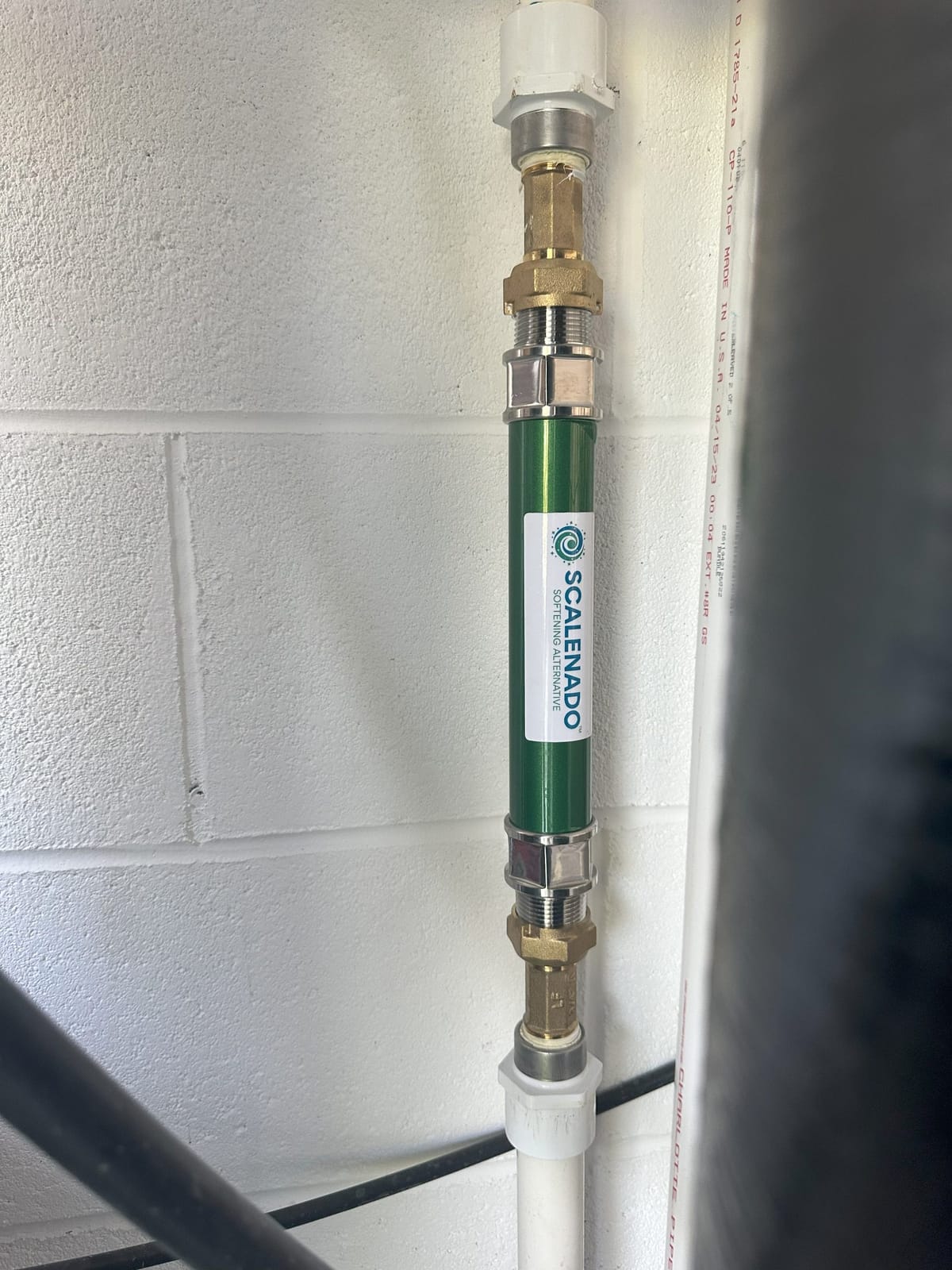 Green Scale Reduction Device Installed on Vertical Water Pipe Against White Brick Wall