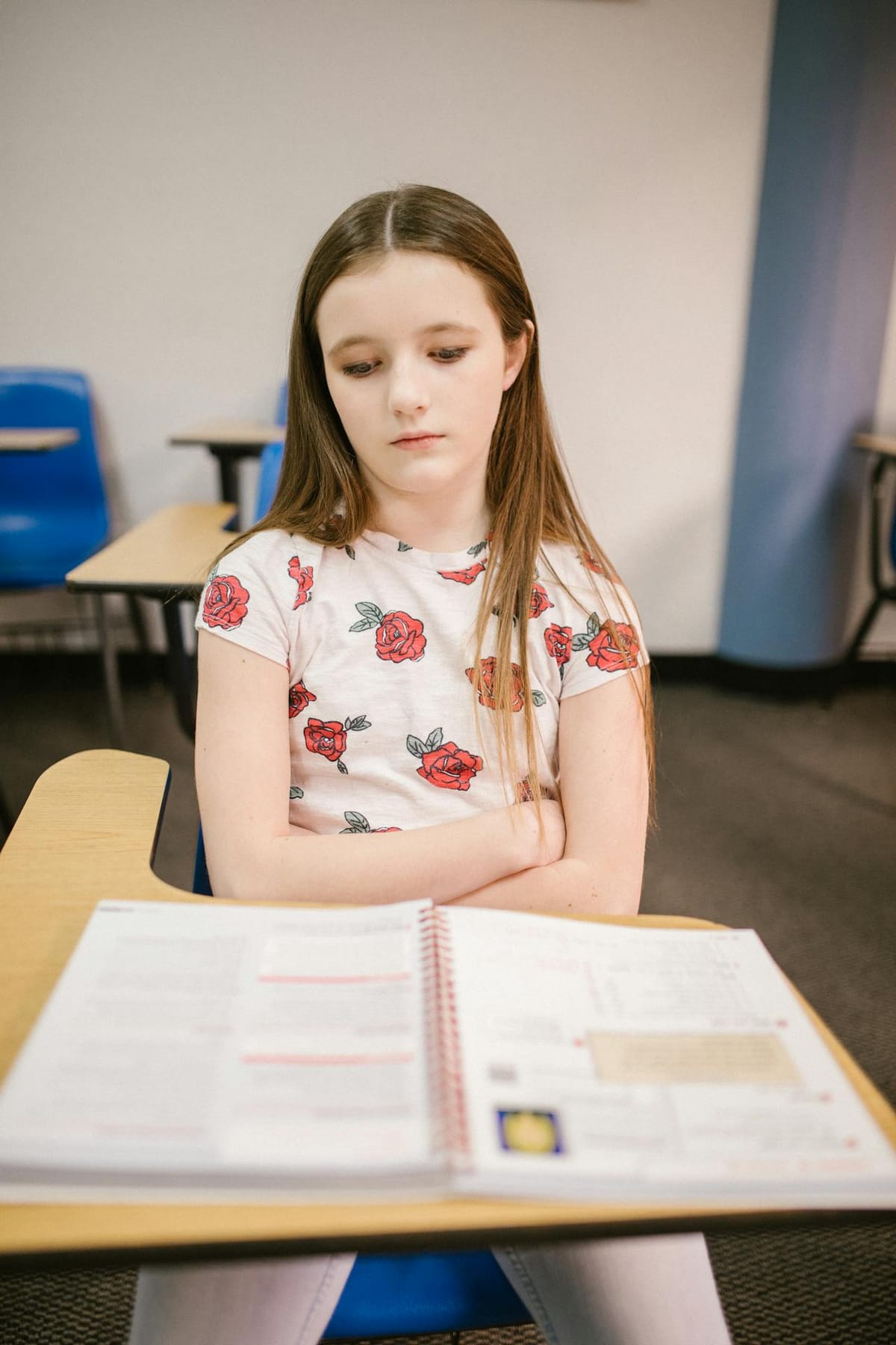 A young girl with long brown hair sits at a school desk in a classroom. She is wearing a white shirt with red rose patterns and appears to be looking down at an open notebook in front of her. The desk is wooden with a blue chair. The classroom has a neutral color scheme with blue accents and additional desks and chairs visible in the background.