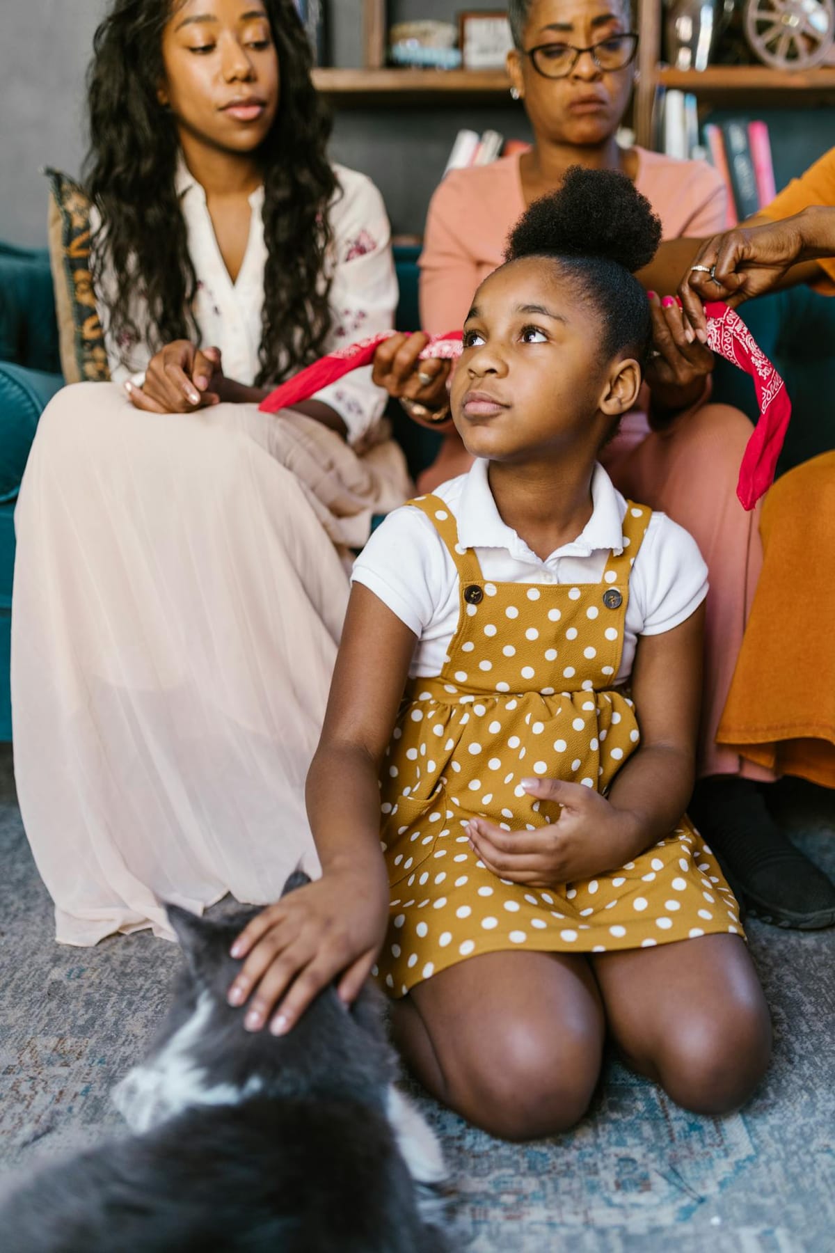 A young girl in a yellow polka dot dress sits on the floor, gently petting a gray and white cat. She is surrounded by three adults, two of whom are tying a red bandana around her head. The scene is set in a cozy living room with a bookshelf in the background. The adults are seated on a teal sofa, and the atmosphere is warm and familial.