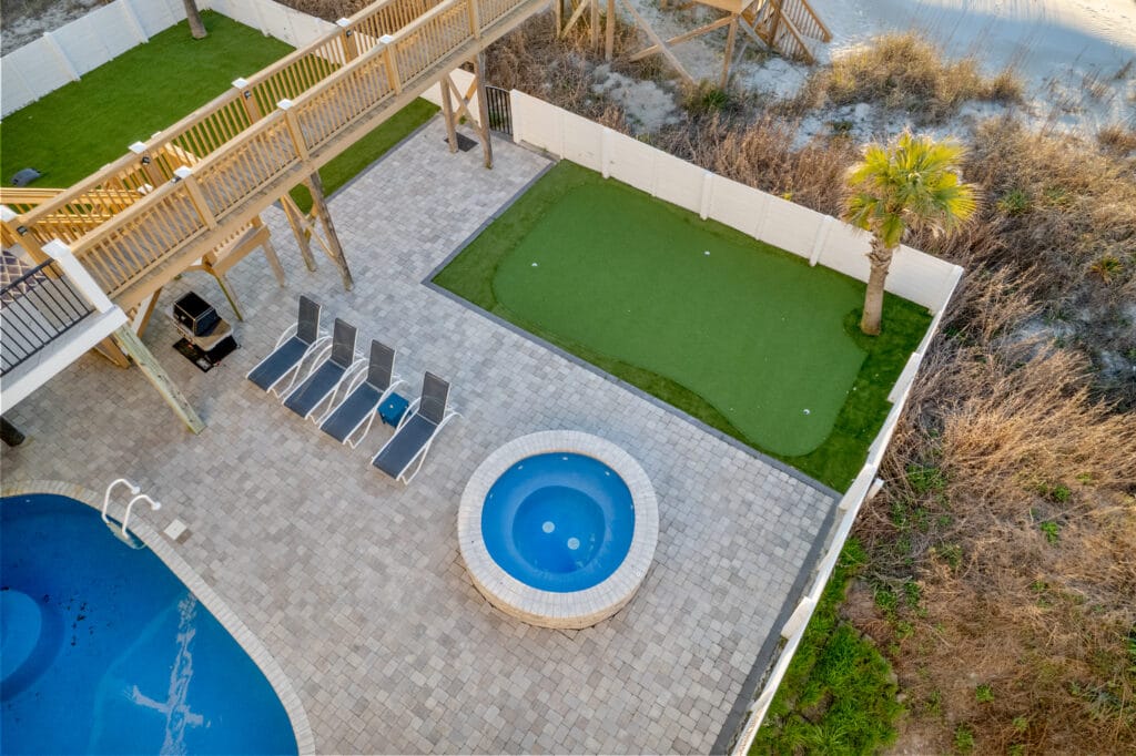 Aerial View of Luxurious Backyard with Pool, Hot Tub, Sun Loungers, Wooden Walkway, and Putting Green Surrounded by White Fence and Lush Vegetation