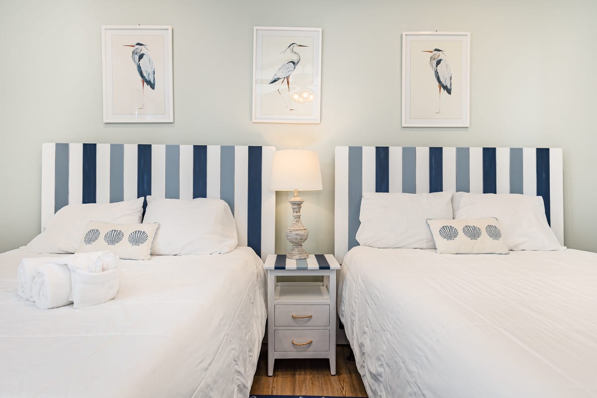 Coastal-Themed Bedroom with Twin Beds Featuring Blue and White Striped Headboards, Central Nightstand with Lamp, and Nautical Bird Artwork on Light Walls