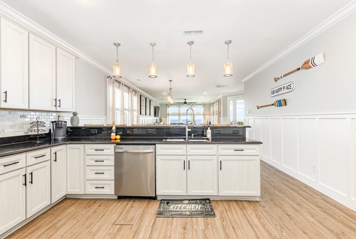 Modern Coastal Kitchen with White Cabinets, Black Countertops, and Nautical Decor Featuring Hanging Pendant Lights, Stainless Steel Appliances, and Wooden Floorboards in Bright Open-Plan Space