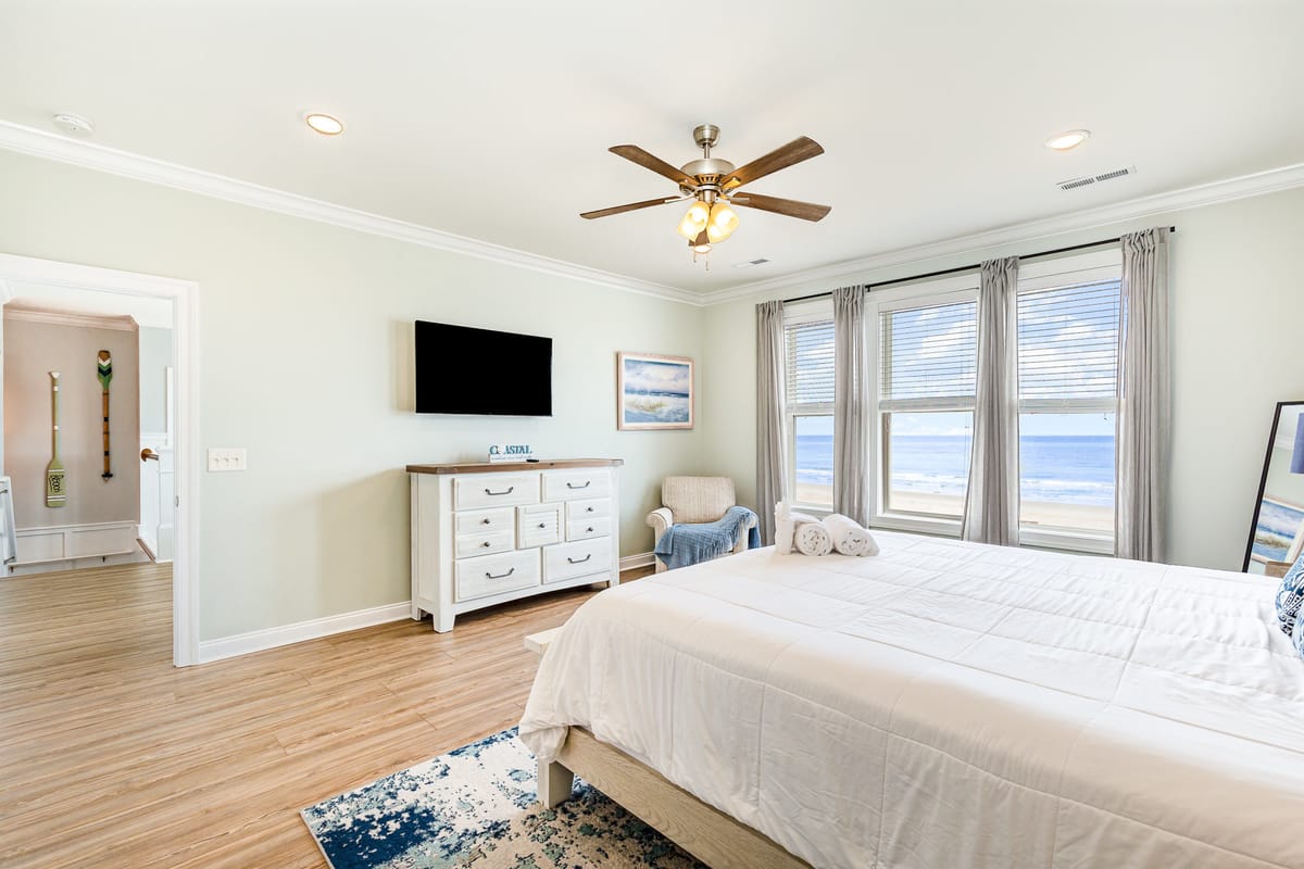 Coastal-Inspired Bedroom with Ocean View, Modern Ceiling Fan, Wall-Mounted TV, and Elegant White Furniture Featuring Decorative Art and Soft Textiles