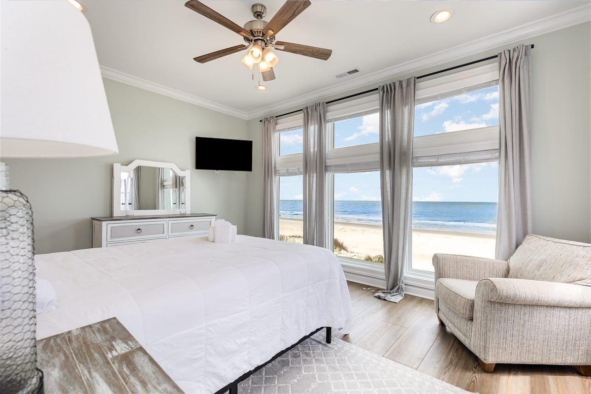 Coastal Bedroom Retreat with Ocean View, White Bedding, and Modern Decor Featuring Ceiling Fan, Light Gray Curtains, and Plush Armchair