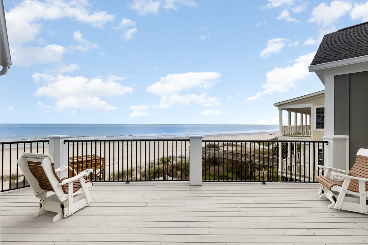 Scenic Coastal View from Wooden Deck with Adirondack Chairs Overlooking Sandy Beach and Ocean Under Blue Sky with Fluffy Clouds
