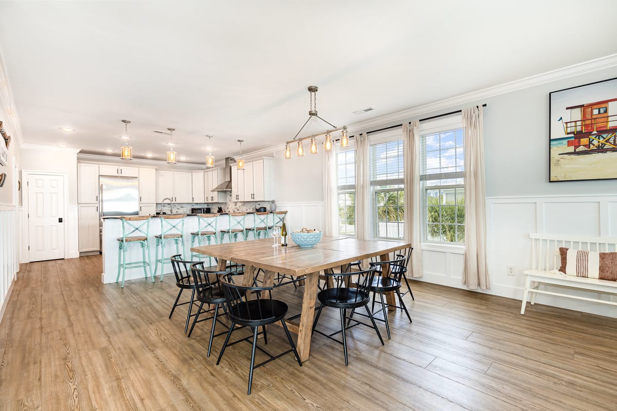 Spacious Open-Concept Dining and Kitchen Area with Wooden Floors, Large Rustic Dining Table, Black Chairs, Modern White Cabinetry, Stainless Steel Appliances, and Coastal Artwork in Sunlit Room with Large Windows and Pendant Lighting.