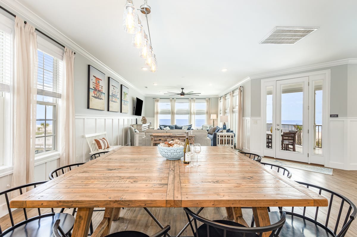 Spacious Coastal Dining and Living Area with Wooden Table, Modern Lighting, Beach Artwork, and Ocean View Through French Doors