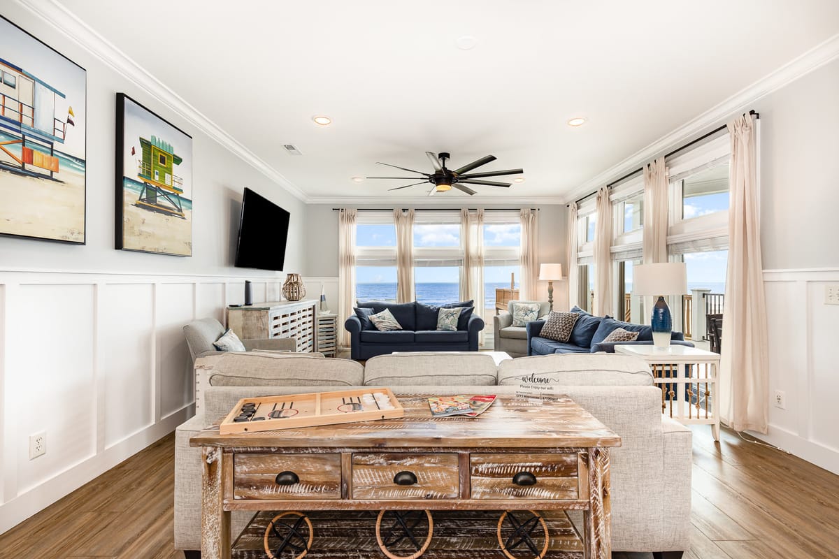 Coastal-Themed Living Room with Ocean View Featuring Blue Sofas, Rustic Wooden Table, Nautical Artwork, and Abundant Natural Light Through Large Windows