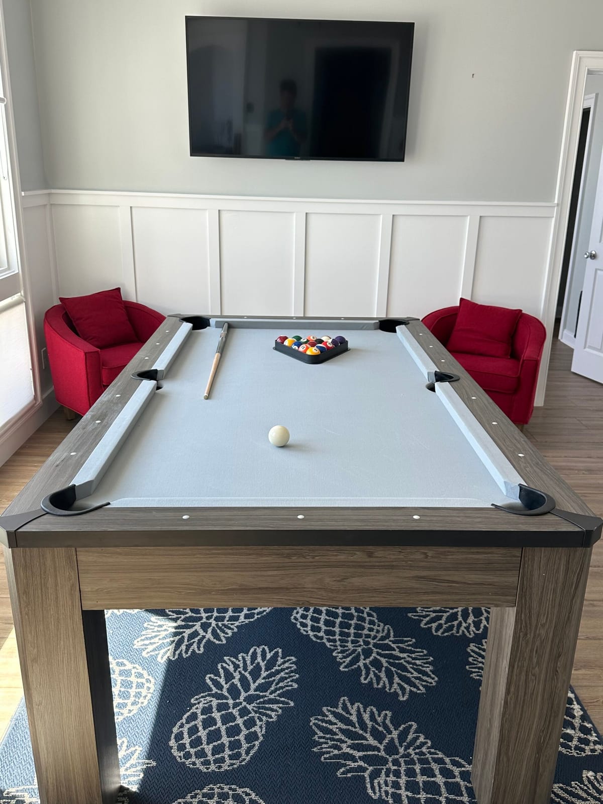 Modern Billiard Room with Light Wood Pool Table, Red Armchairs, Large Wall-Mounted TV, and Pineapple Patterned Rug