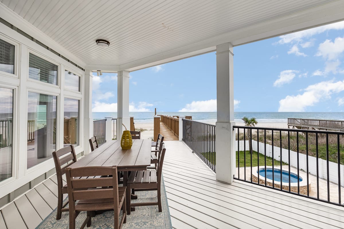 Spacious Oceanfront Patio with Wooden Dining Set Overlooking Beach and Jacuzzi, Framed by White Railings and Bright Blue Sky