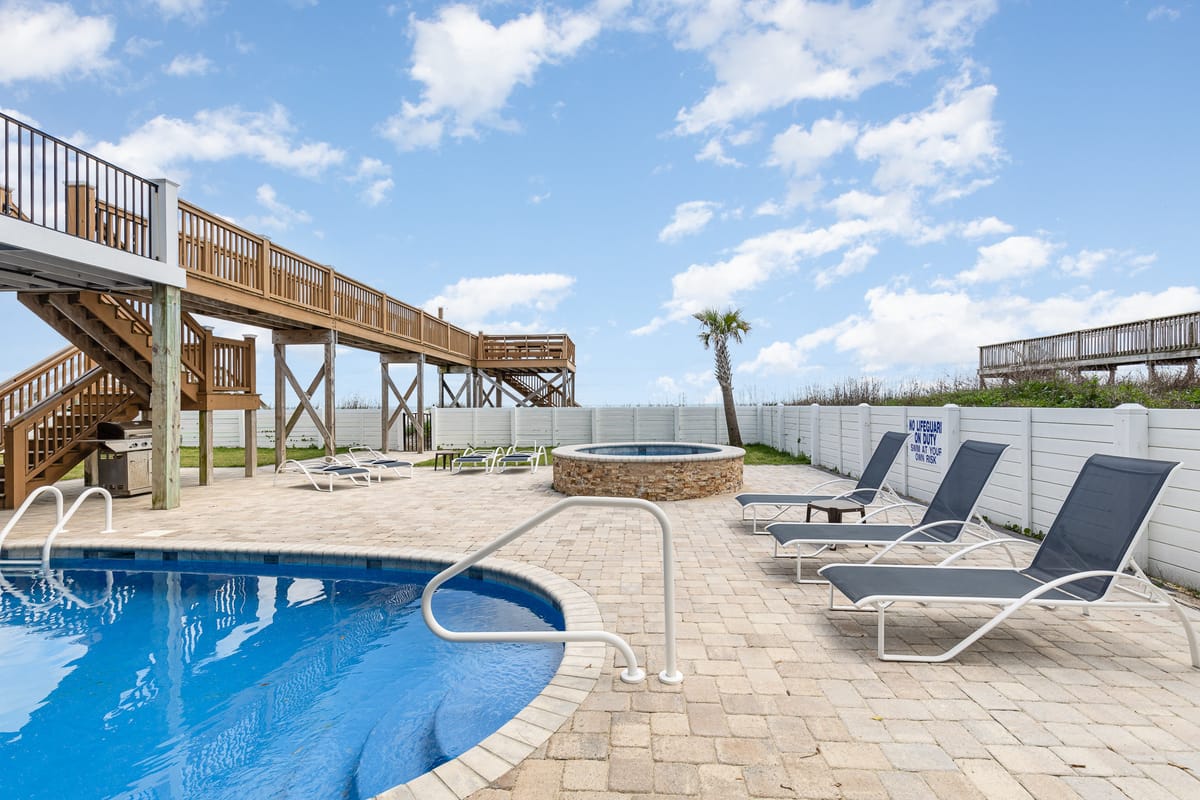 Spacious Outdoor Pool Area with Curved Blue Pool, Stone Hot Tub, Sun Loungers, and Elevated Wooden Walkway Under Bright Blue Sky with Fluffy White Clouds