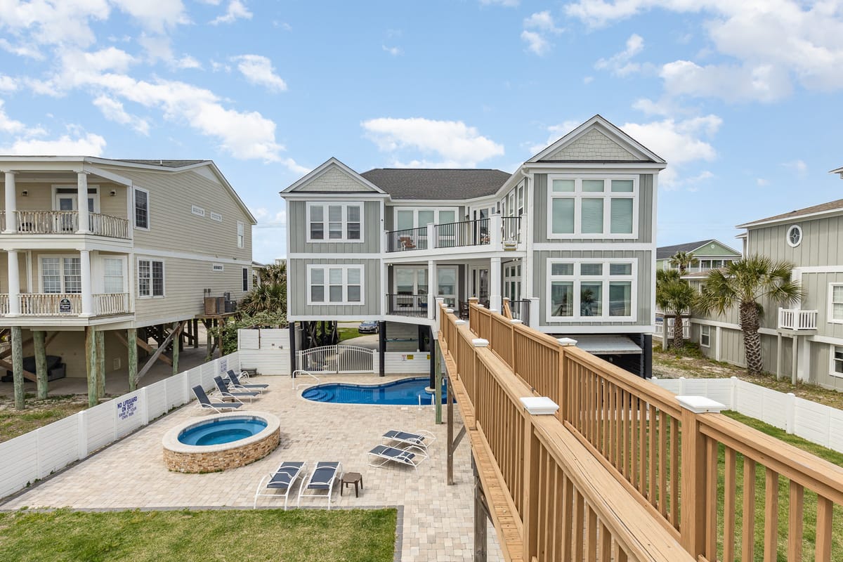 Spacious Coastal Beach House with Elevated Wooden Walkway, Outdoor Pool and Hot Tub Surrounded by Lounge Chairs and Palm Trees Under a Clear Blue Sky