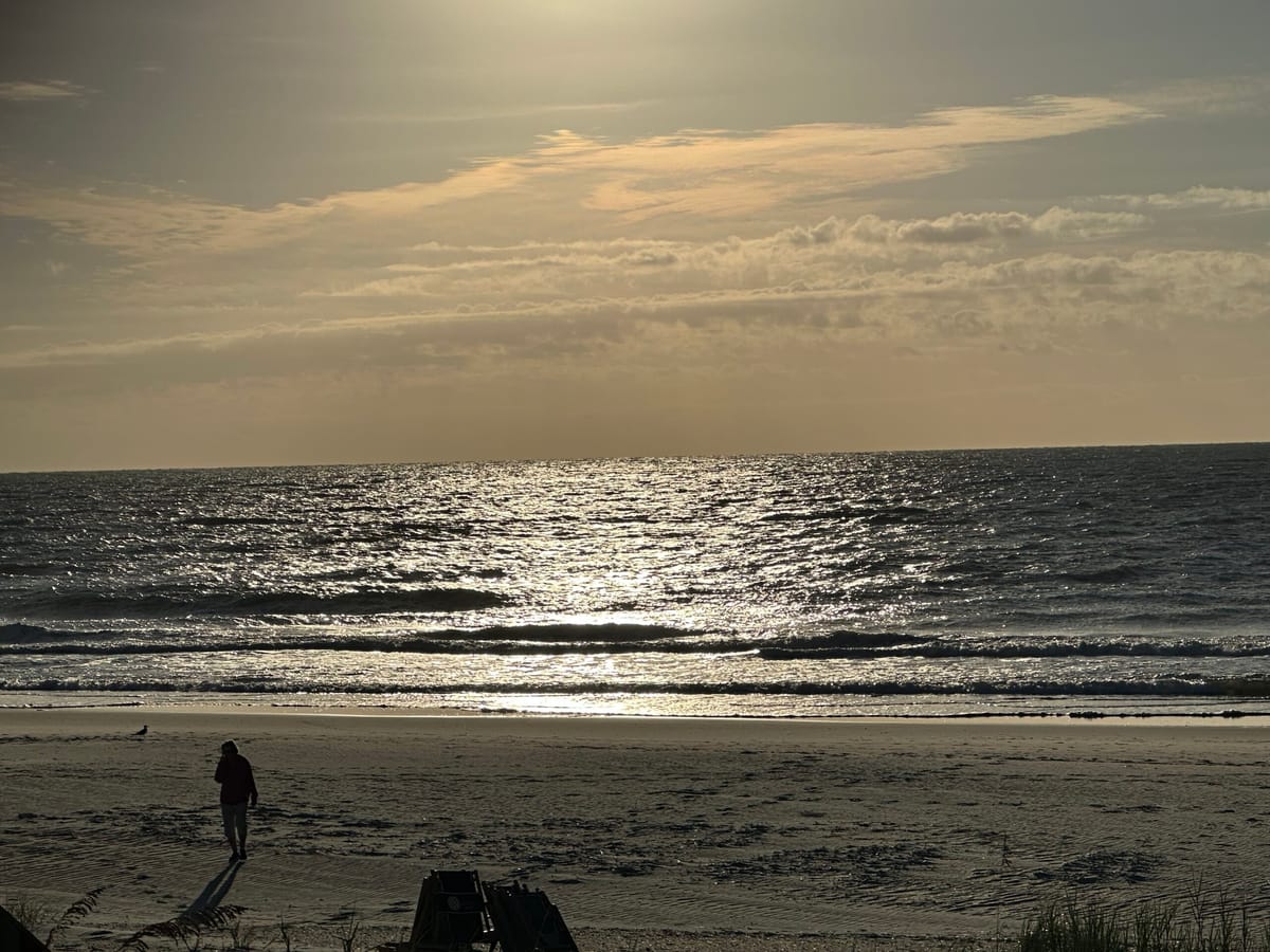 Solitary Figure Walking Along Tranquil Beach at Sunrise with Reflective Ocean Waves and Golden Sky