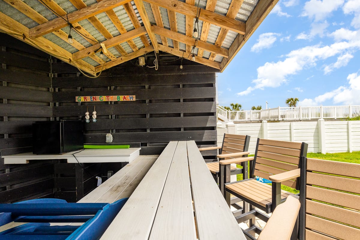 Rustic Outdoor Patio with Wooden Seating and Table Under a Wooden Roof, Featuring String Lights and a Decorative Sign, Overlooking a Grassy Area with a White Fence and Clear Blue Sky